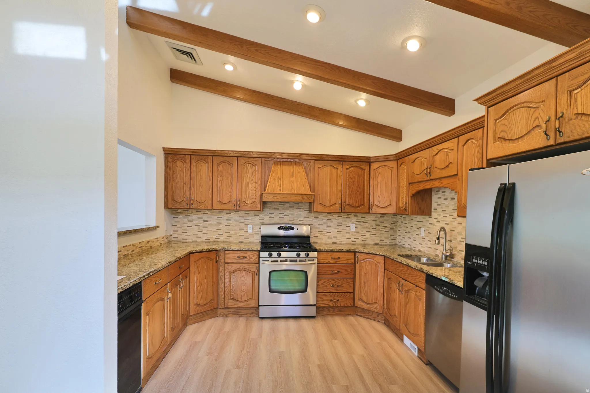 Kitchen featuring wood-finish cabinetry, granite-style countertops, stainless steel appliances, a tiled backsplash, and exposed ceiling beams