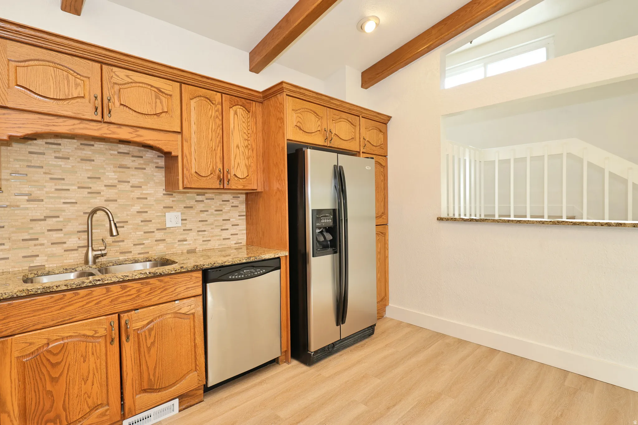 Kitchen featuring a dual basin sink, stainless steel appliances, wood-finish cabinetry, tile backsplash, and exposed ceiling beams