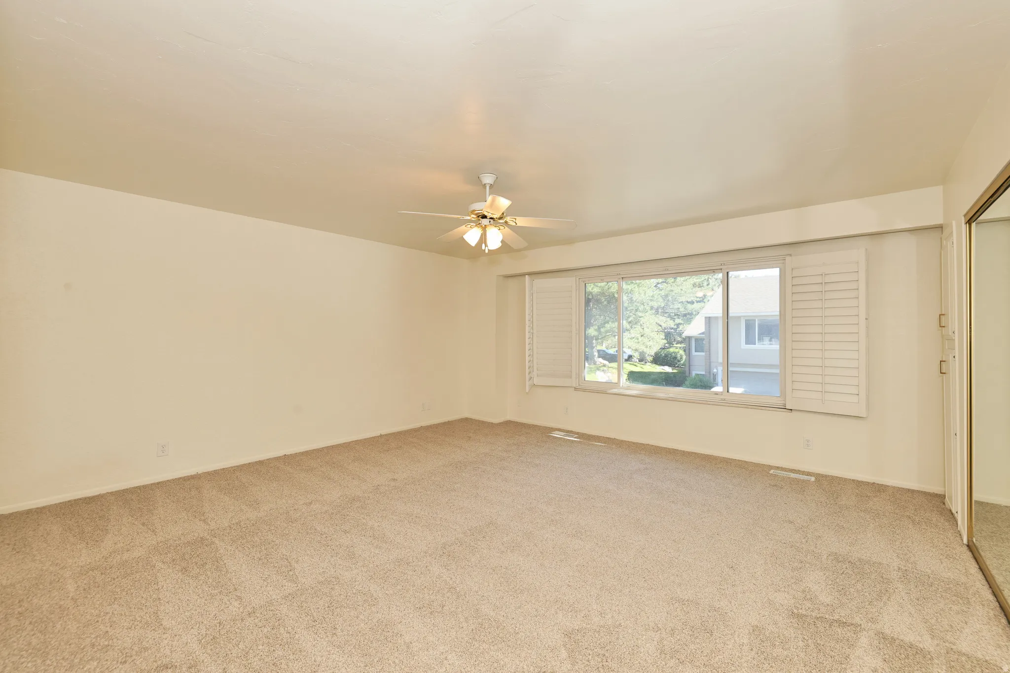 Expansive carpeted room featuring a ceiling fan, a large window with white shutters, and a mirrored closet door