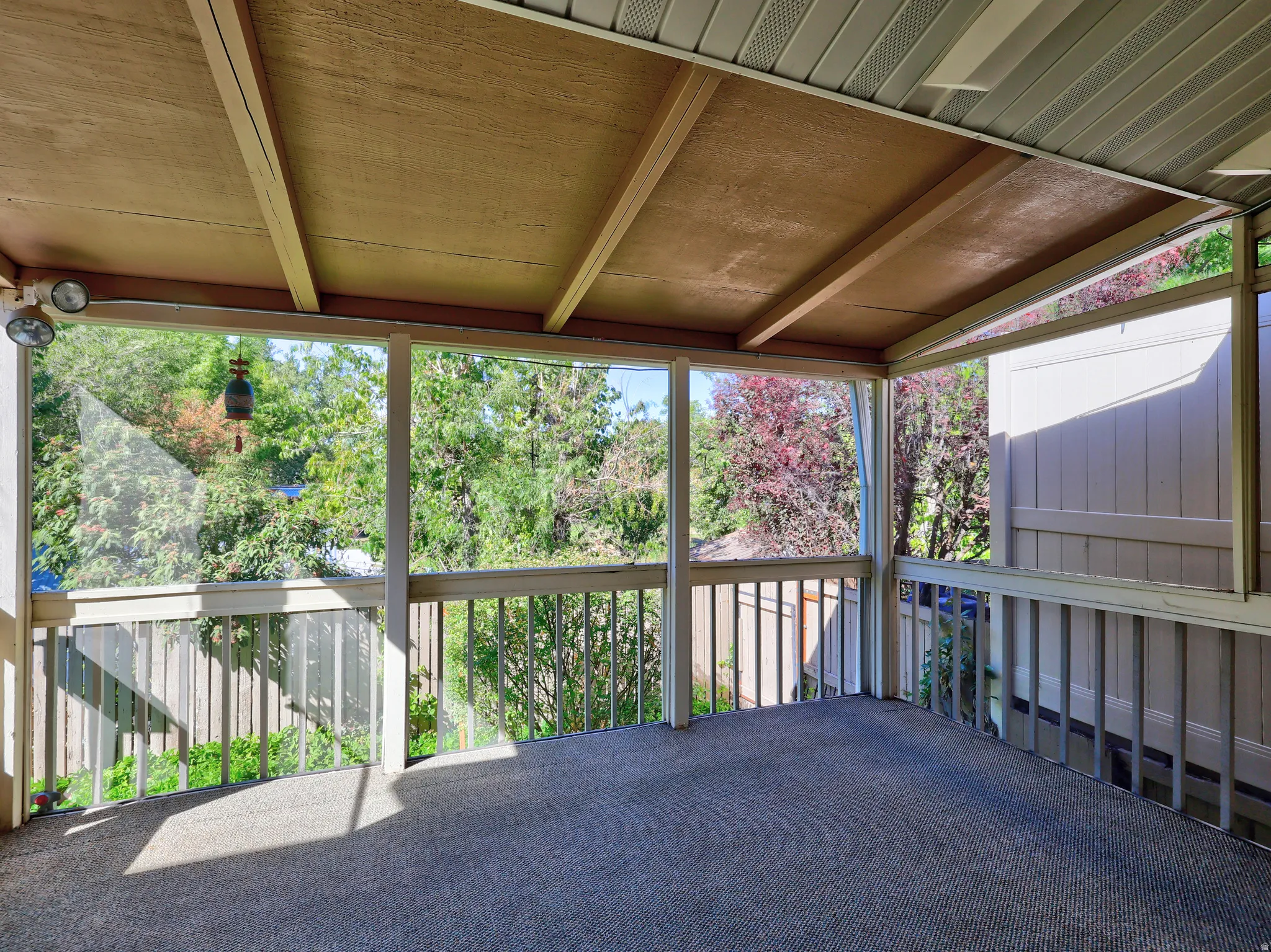 Covered outdoor patio with a carpeted floor, wood railings, and exterior lighting fixtures