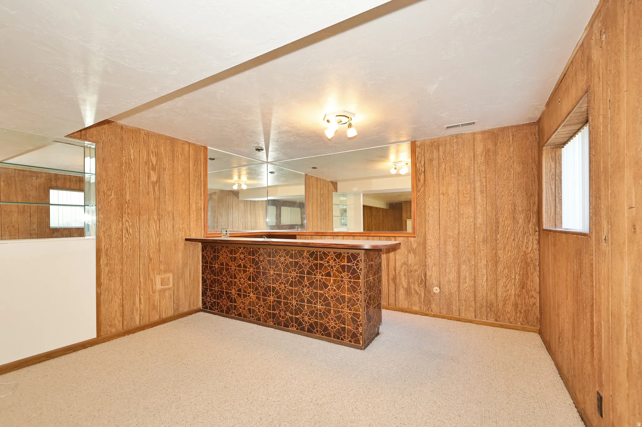 Wood paneling walls, built-in bar with decorative front, mirrored wall accents, light-colored carpeting, and a recessed window