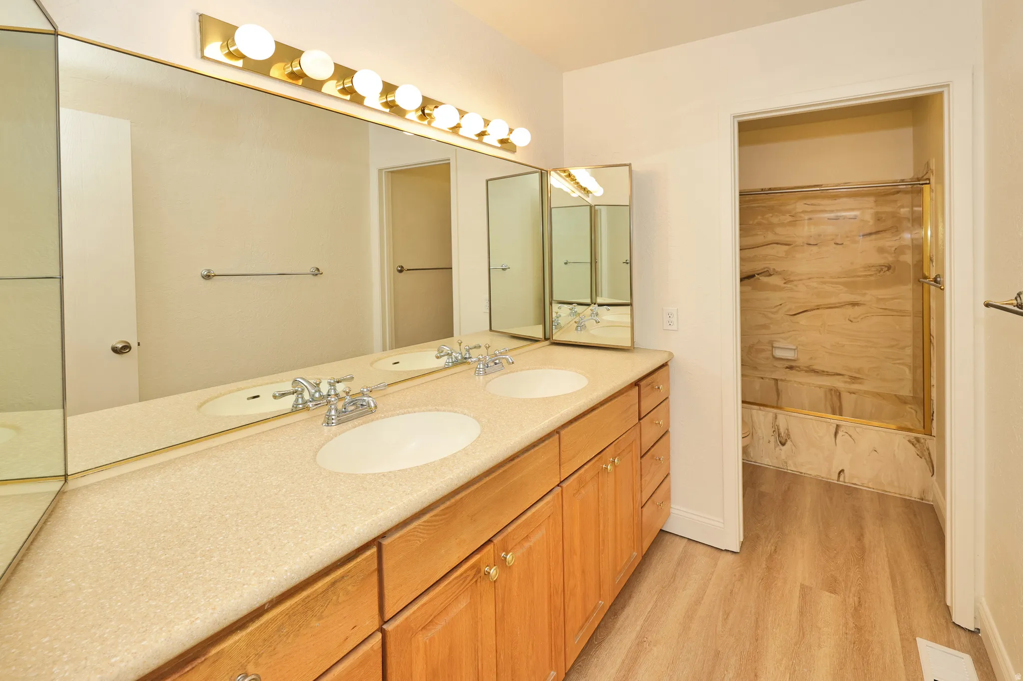 Double vanity with dual sinks, laminate wood-finish flooring, full-width mirror, light wood-finish cabinetry, and a tiled shower-tub combination