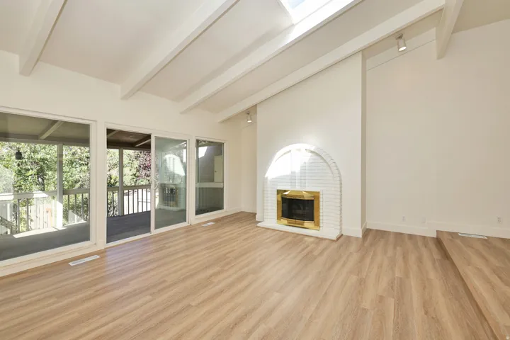 Vaulted ceiling with exposed beams, wood-finish flooring, white brick fireplace with brass surround, multiple glass sliding doors, and a skylight