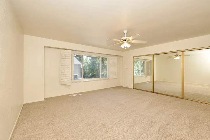 Carpeted room featuring a ceiling fan with light fixture, large windows with white shutters, and mirrored sliding closet doors