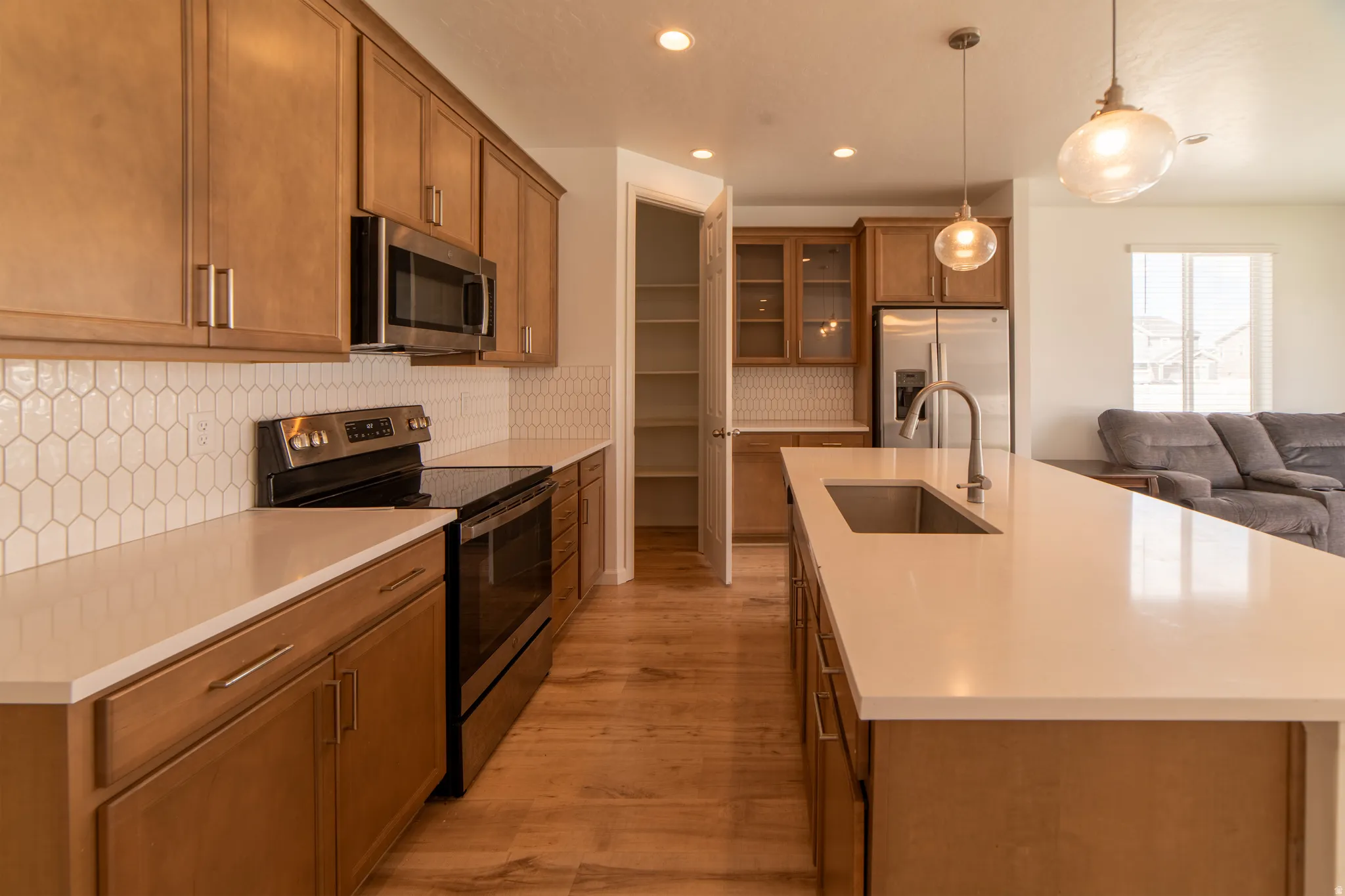 Kitchen featuring wood-finish cabinetry, stainless steel appliances, light countertops, a kitchen island with an undermount sink, and pendant lighting