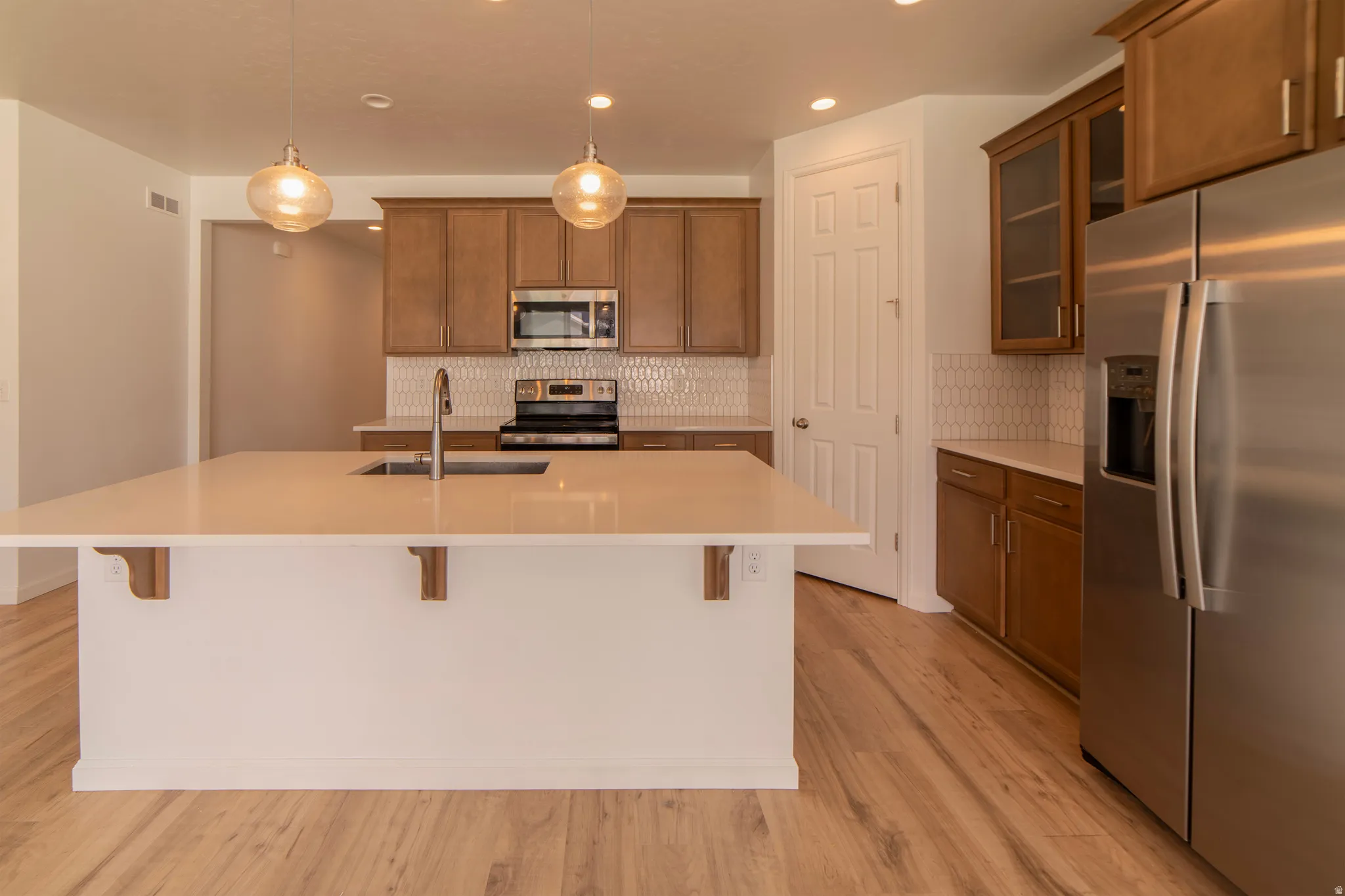 Kitchen featuring an island with an undermount sink, pendant lighting, wood-finish flooring, stainless steel appliances, and a tiled backsplash