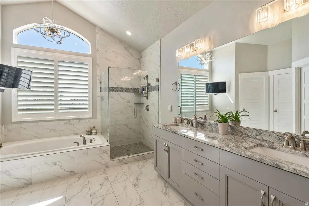 Primary Bathroom- Vaulted ceiling with an arched window, plantation shutters, a glass-enclosed shower, a built-in tub, and a dual vanity with a stone countertop