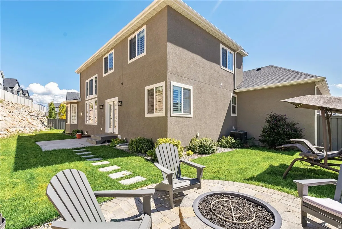Modern home with a spacious backyard featuring a manicured lawn, stone fire pit with integrated seating area, mature landscaping, and a patio extension