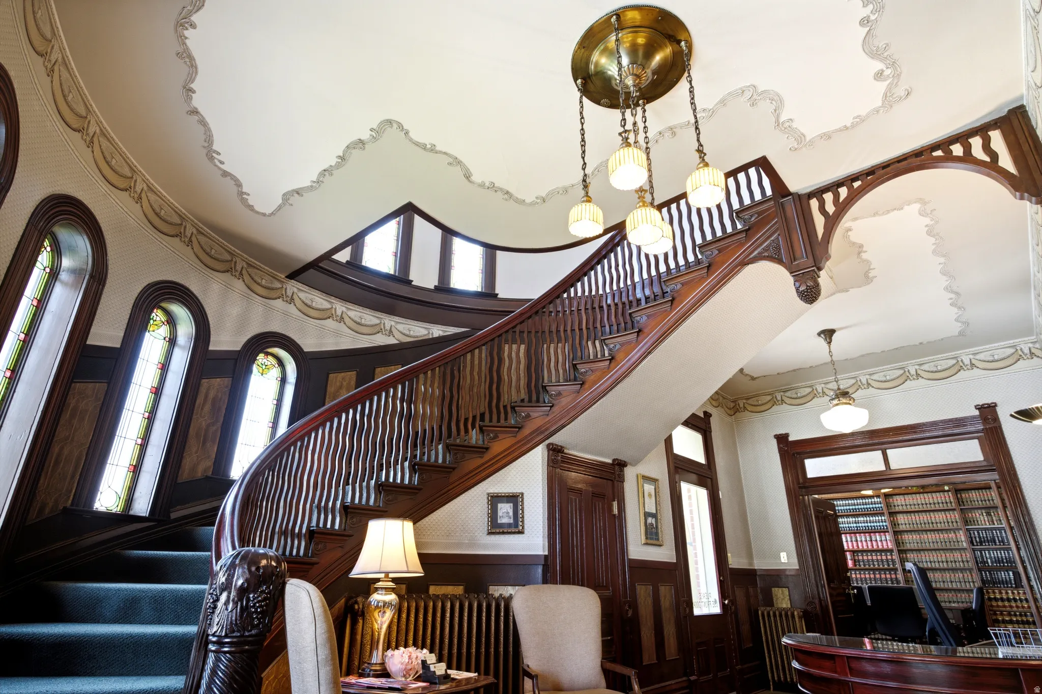Grand curved staircase with ornate wood newel post and balusters, featuring multiple arched stained-glass windows, decorative ceiling trim, and a prominent brass chandelier