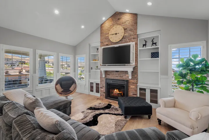 Vaulted ceiling with recessed lighting, brick fireplace with mantel, built-in shelving, wood-finish flooring, and multiple windows with plantation shutters