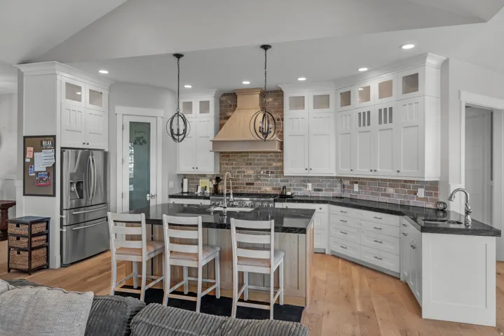 White shaker-style cabinetry with upper glass-front accents, dark countertops, brick-finish backsplash, and wood-finish flooring