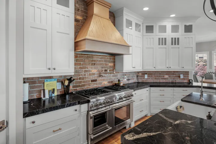 White shaker-style cabinetry with upper glass-front cabinets, brick backsplash, dark countertops with light veining, stainless steel range with double oven, and wood-finish range hood