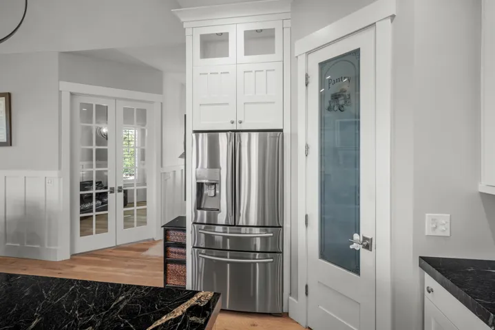 Stainless steel refrigerator, white shaker-style cabinetry, glass-panel pantry door, black countertop, and wood-finish flooring