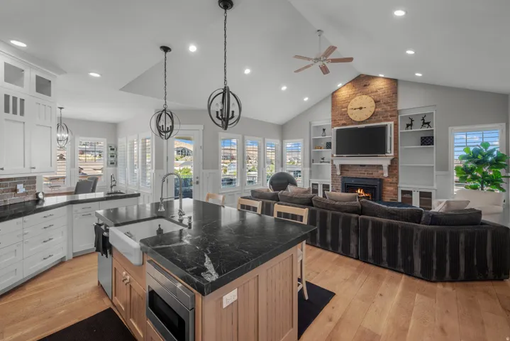 Open concept living space featuring vaulted ceilings, wood-finish flooring, a kitchen island with an integrated farmhouse sink, white cabinetry, and a brick fireplace