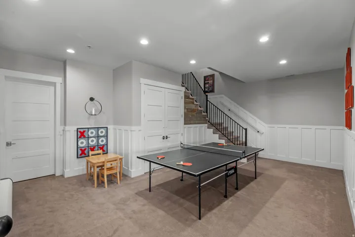 Recessed lighting illuminates a carpeted room featuring white panel wainscoting and a staircase with a black metal railing