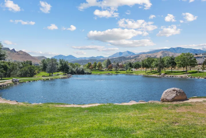 Lush green foreground with a tranquil pond, bordered by a walking path and mature trees, set against a backdrop of distant mountains