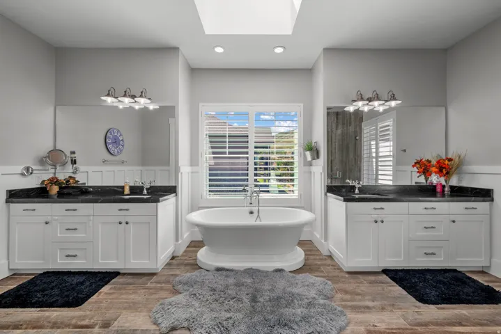 Dual white vanities with dark countertops, a central freestanding bathtub, wood-finish flooring, and a skylight