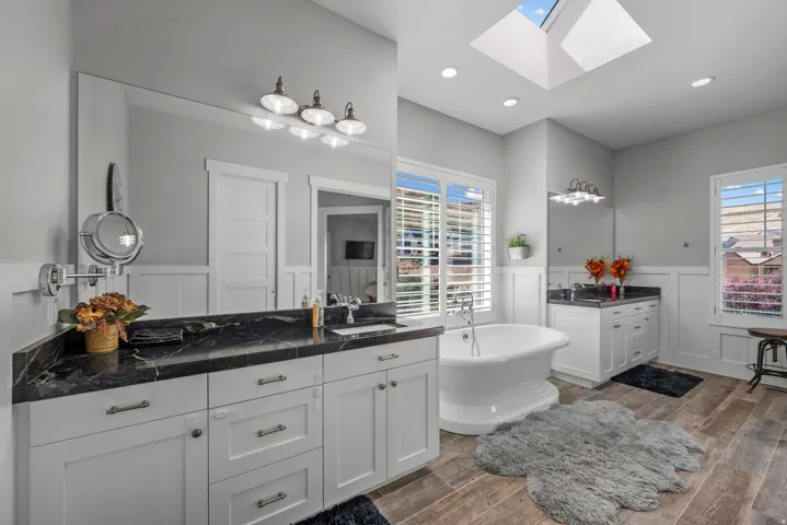 Freestanding soaking tub, wood-finish flooring, dual vanity with dark stone countertops, skylight, and white paneling