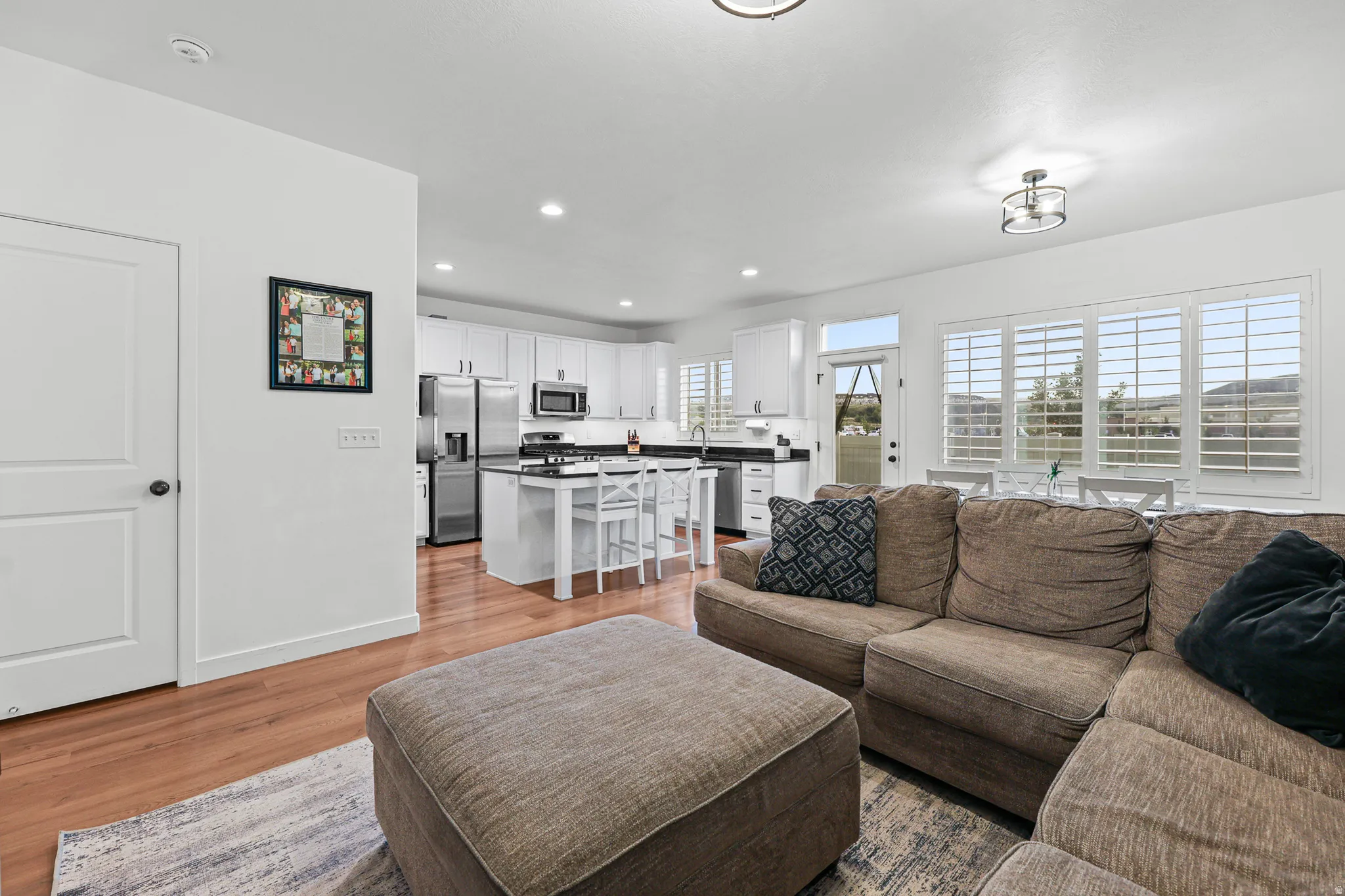 Open-concept living space featuring wood-finish flooring, white kitchen cabinetry, stainless steel appliances, a kitchen island with seating, and plantation shutters