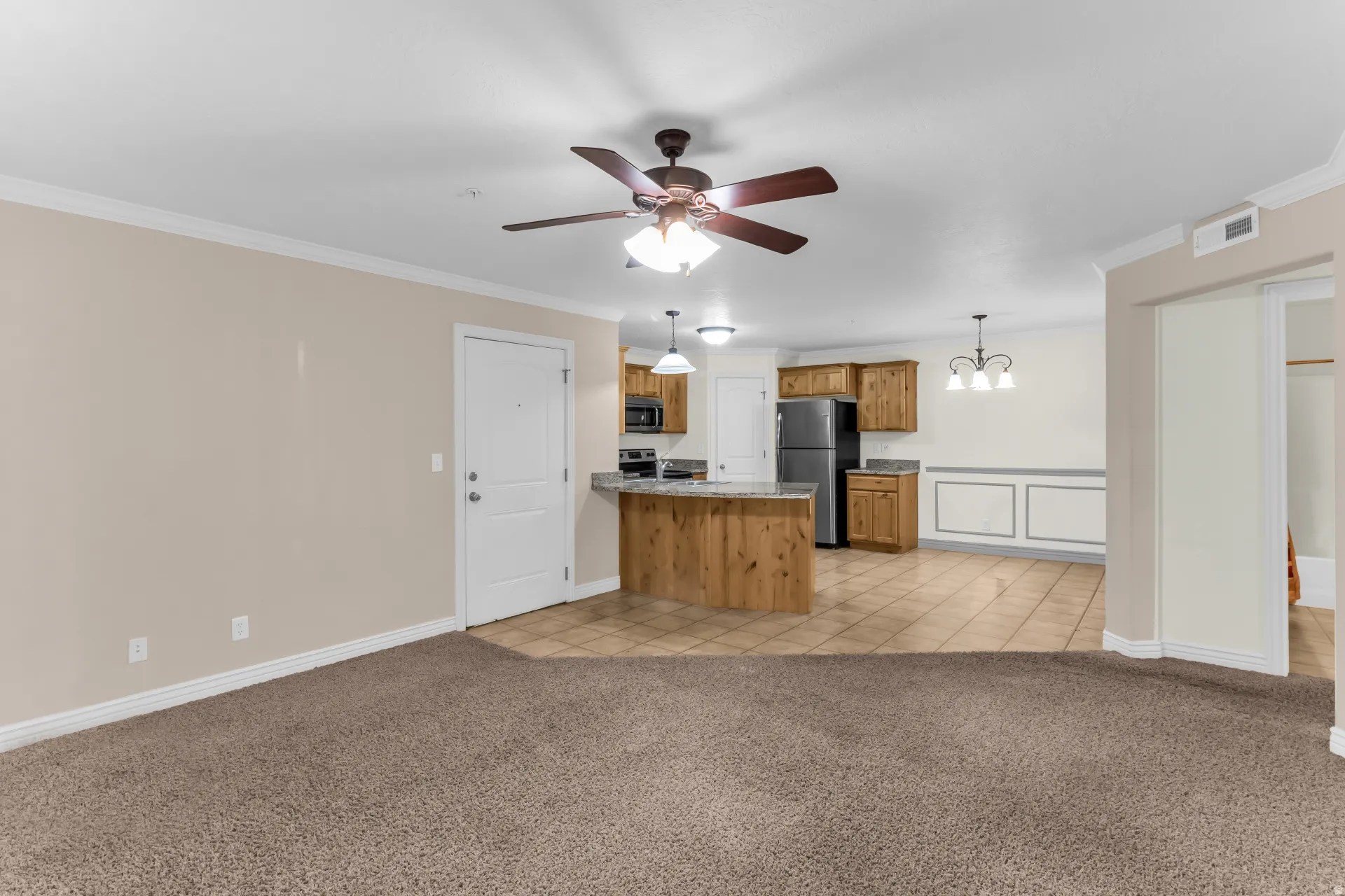 Carpeted living area with crown molding, a ceiling fan, and a kitchen featuring wood cabinetry, stainless steel appliances, and a breakfast bar