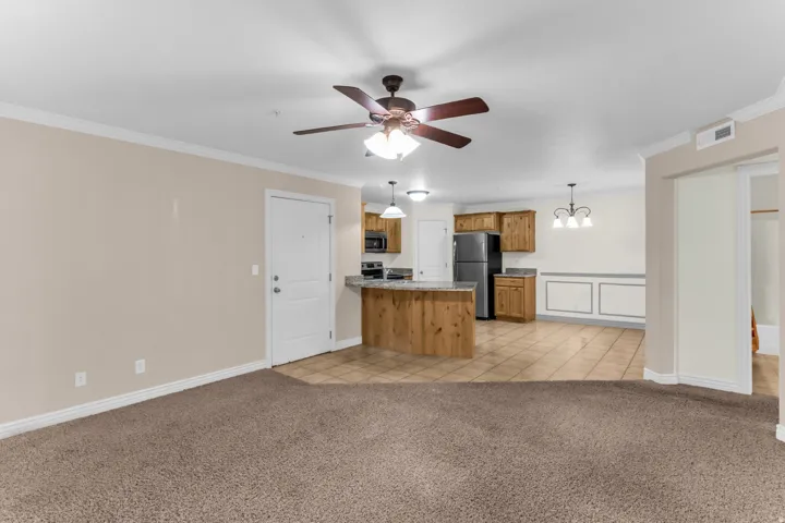 Carpeted living area with crown molding, a ceiling fan, and a kitchen featuring wood cabinetry, stainless steel appliances, and a breakfast bar