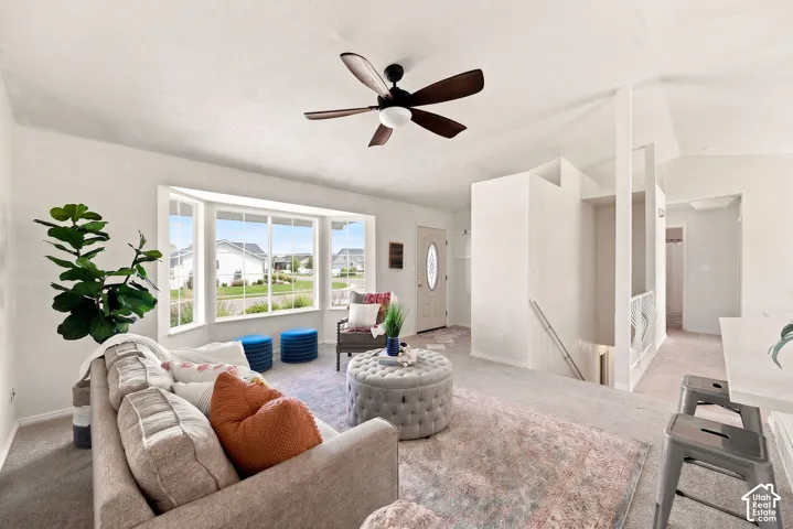 Spacious living area featuring a bay window, ceiling fan with light fixture, light-toned carpeting, and a built-in staircase