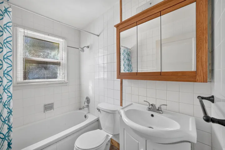 Bathroom featuring white tile walls, a white bathtub with shower fixture, a window with blinds, a wood-finish medicine cabinet, and a vanity with an integrated sink
