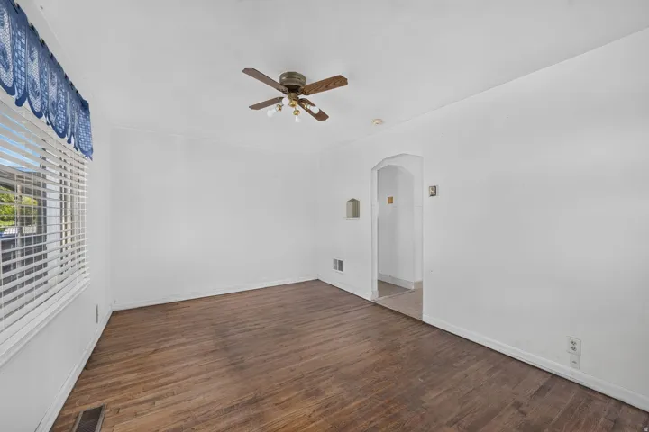 Wood-finish flooring, white walls, ceiling fan, window with blinds, and an arched doorway