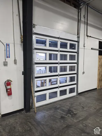 White overhead door with multiple window panels, industrial concrete flooring, exposed overhead piping, and a metal support beam