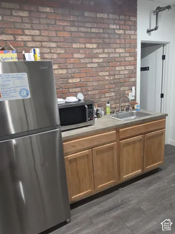 Brick-finish accent wall with wood-finish flooring, stainless steel refrigerator, and wood cabinetry