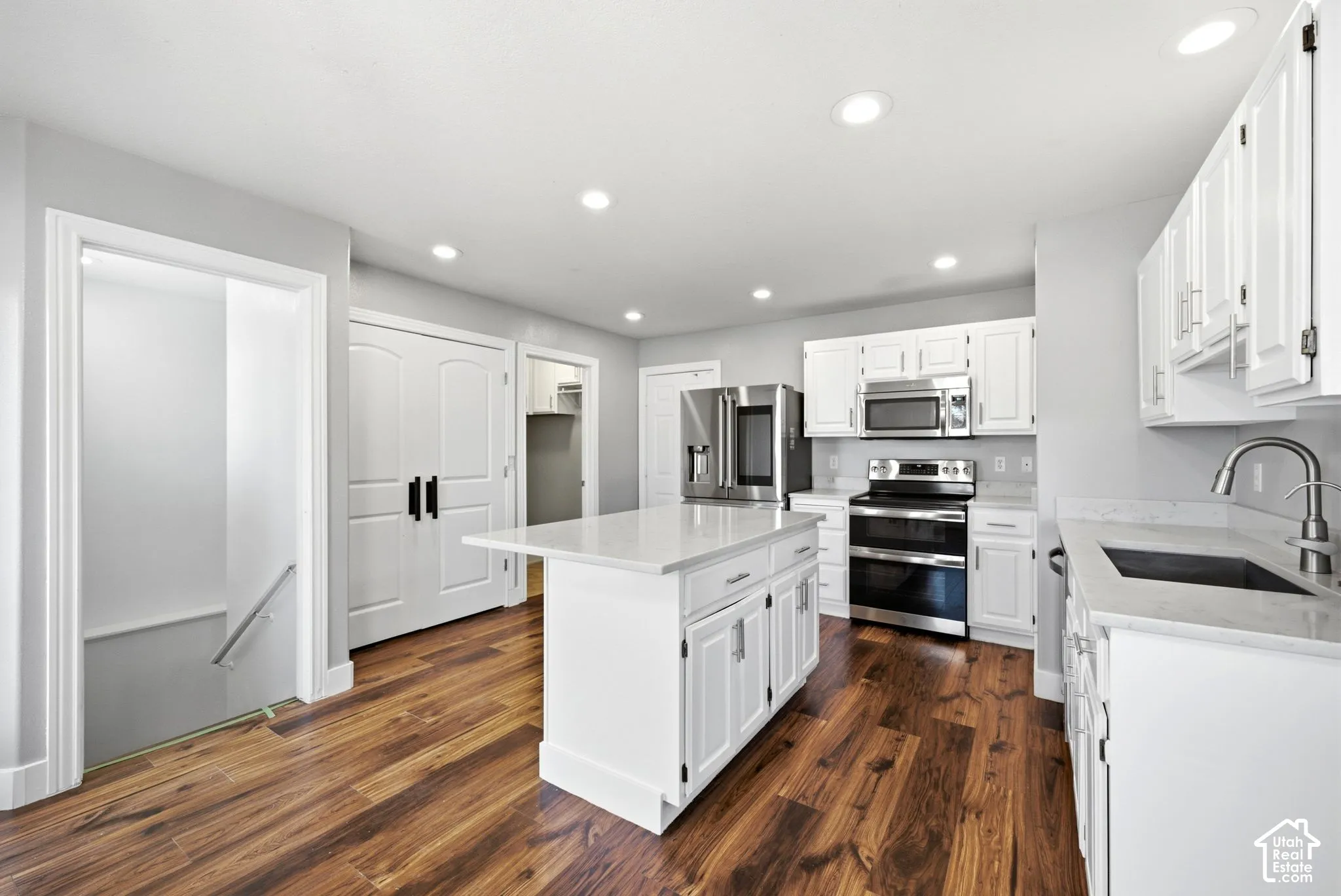 Wood-finish flooring, white cabinetry with silver-tone hardware, stainless steel appliances, recessed ceiling lights, and a white island with a light-toned countertop