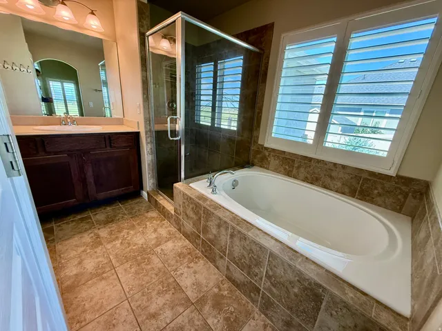 Master Bathroom Tiled flooring, built-in soaking tub, glass-enclosed shower, wood-finish vanity with countertop, and window with plantation shutters