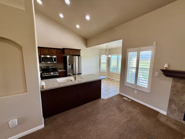 Vaulted ceiling with recessed lighting, kitchen with stainless steel appliances, breakfast bar with integrated sink, tiled fireplace surround, and plantation shutters