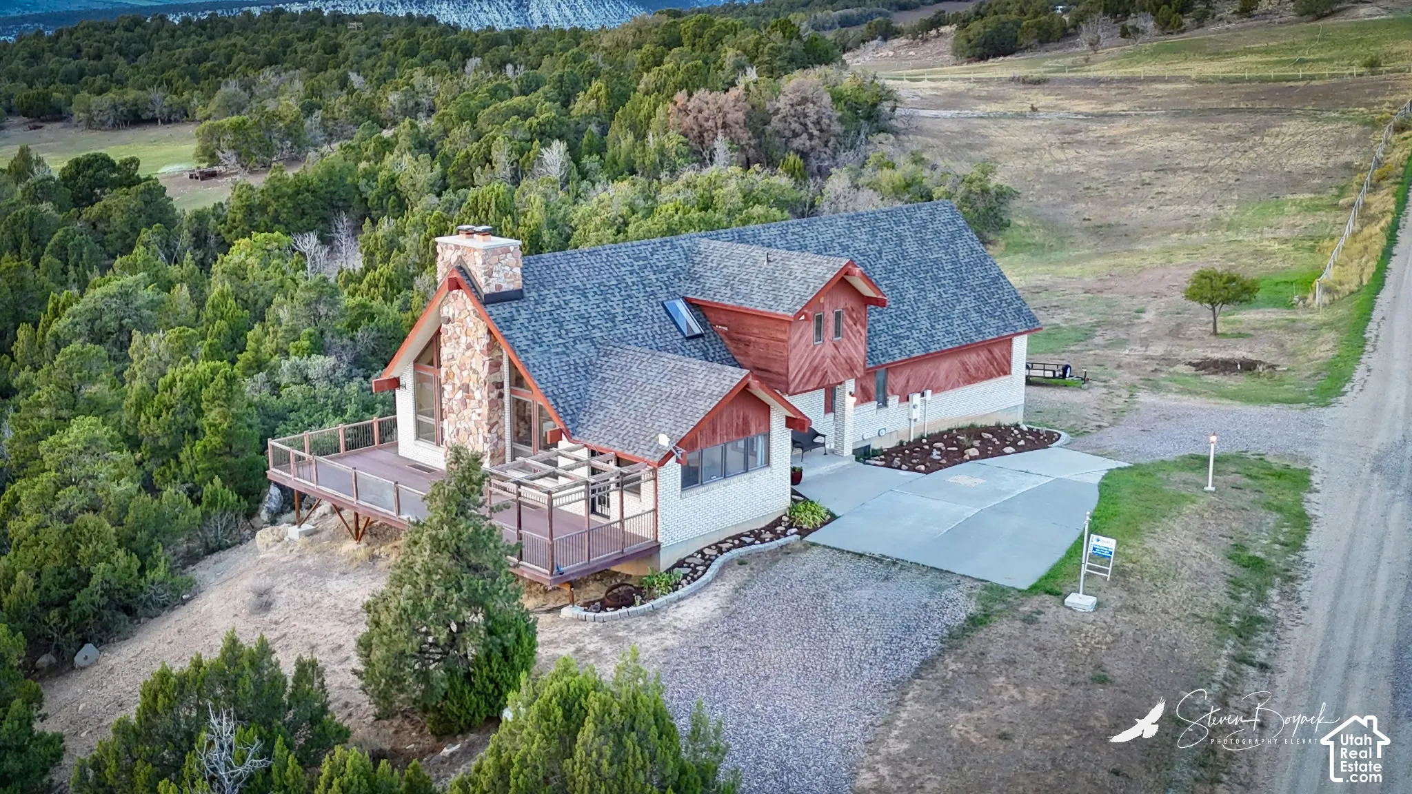Stone-clad chimney exterior with a pitched roof, an expansive outdoor deck, a paved driveway, and mature surrounding landscaping