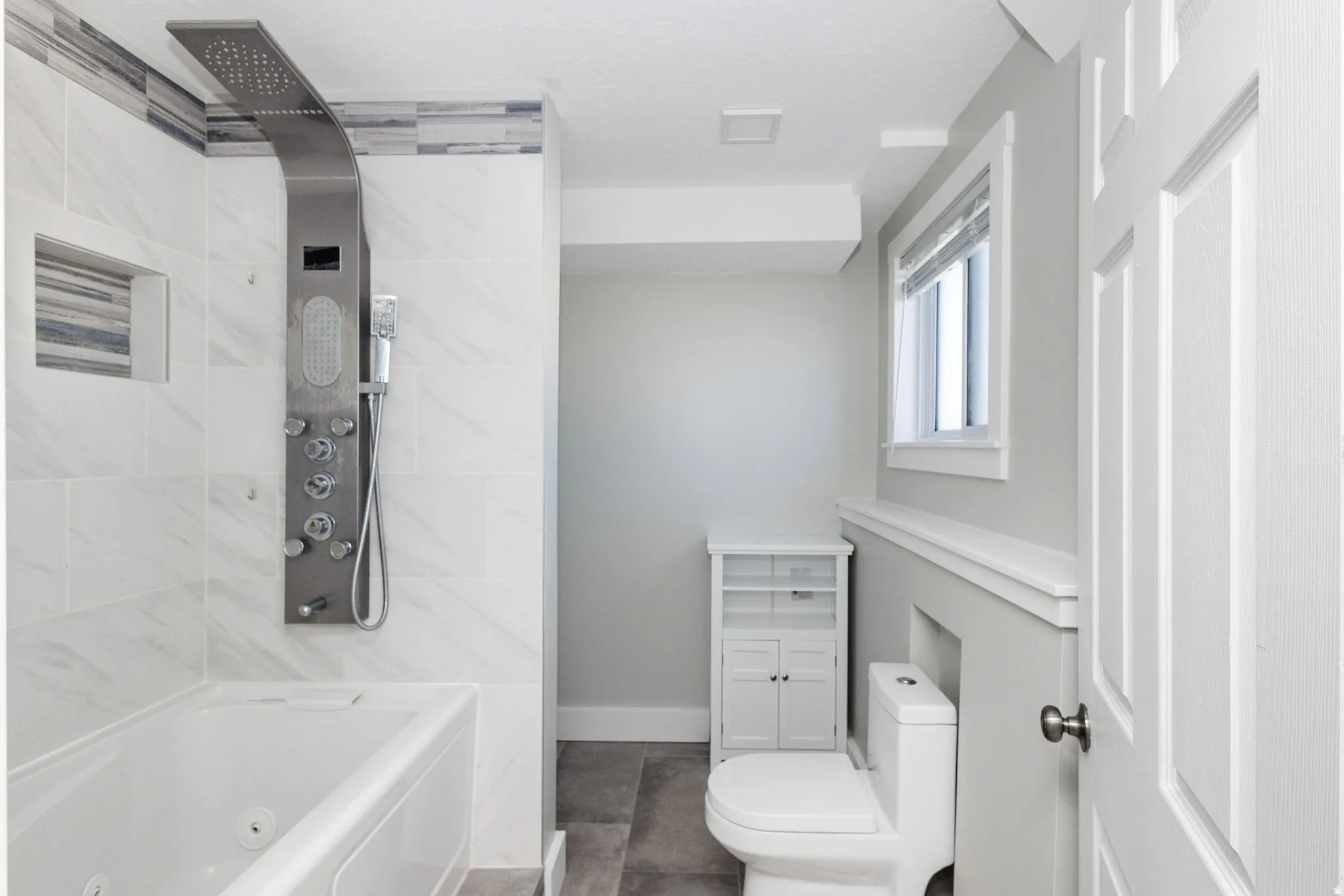 Jetted soaking tub with a multi-function shower panel, white subway tile surround, built-in shower niche, and grey wood-finish flooring