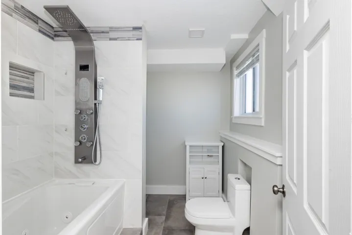 Jetted soaking tub with a multi-function shower panel, white subway tile surround, built-in shower niche, and grey wood-finish flooring