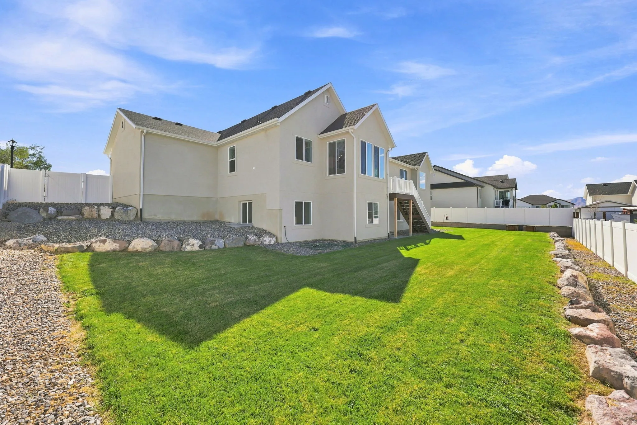 Expansive grassy lawn with a gravel border, featuring a stucco exterior with multiple windows, and a raised exterior deck with stairs