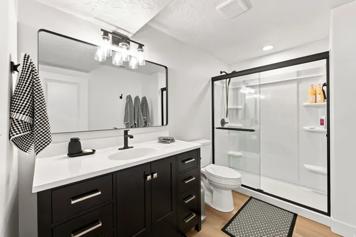Modern vanity with a white countertop and dark cabinetry, large framed mirror, matte black fixtures, and a glass-enclosed shower with a sliding door
