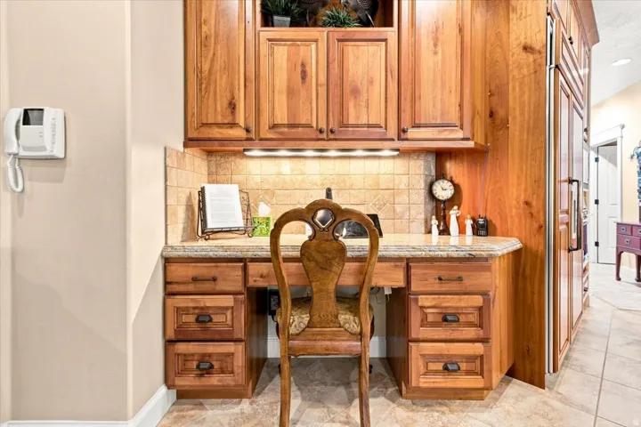 Built-in desk featuring wood cabinetry, a tiled backsplash, under-cabinet lighting, and a stone countertop