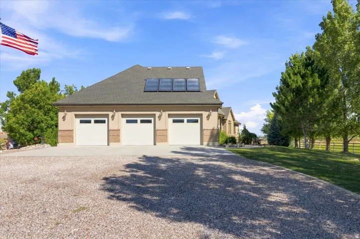 Three-car garage with overhead doors and transom windows, stucco exterior, asphalt shingle roof with solar panels, and an extended gravel driveway