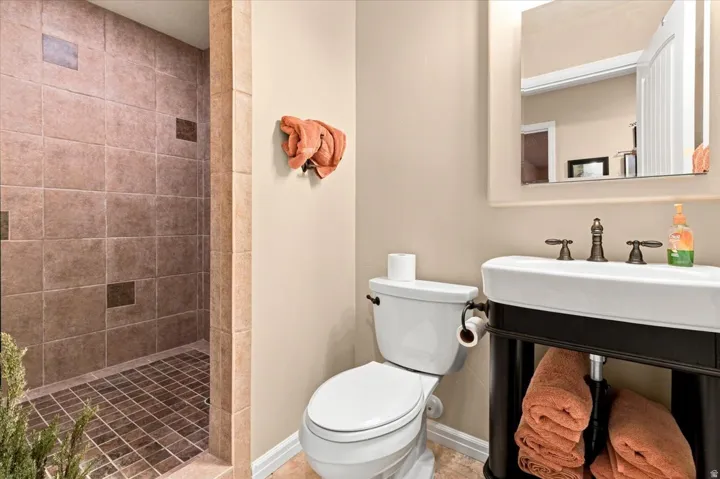 Walk-in shower with tiled walls and floor, pedestal sink with dark bronze fixtures, wall-mounted mirror, and toilet