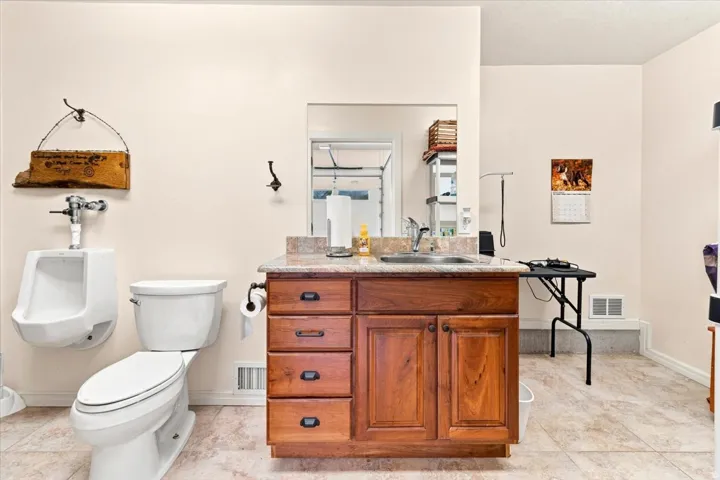 Ceramic tile flooring, white toilet, wall-mounted urinal, wood-finish vanity with stone countertop, and integrated sink