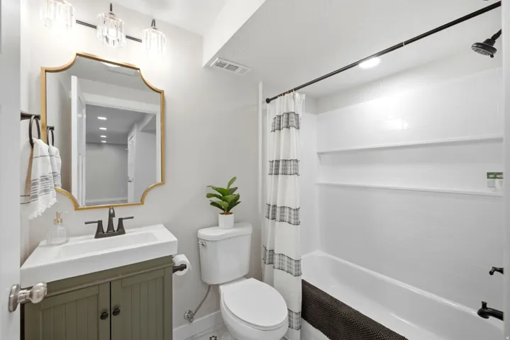 White tiled shower with built-in shelving, a white porcelain tub, a vanity with a white basin and dark green cabinetry, a decorative gold-framed mirror, and a three-light vanity fixture