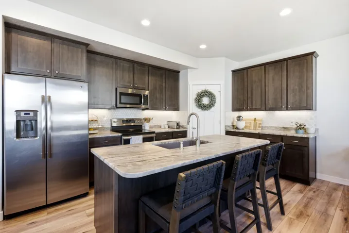 Kitchen featuring wood-finish flooring, dark wood cabinetry, stainless steel appliances, and a central island with a sink and stone countertop