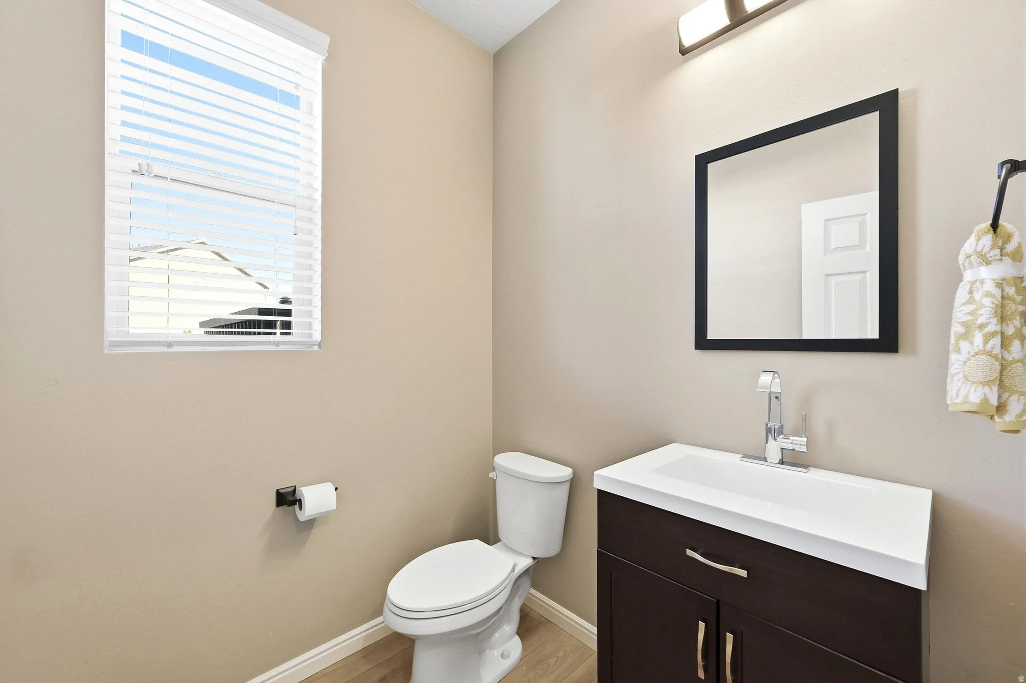 Neutral-toned walls, wood-finish flooring, a dark wood-finish vanity with an integrated white sink, a framed mirror, and a window with blinds
