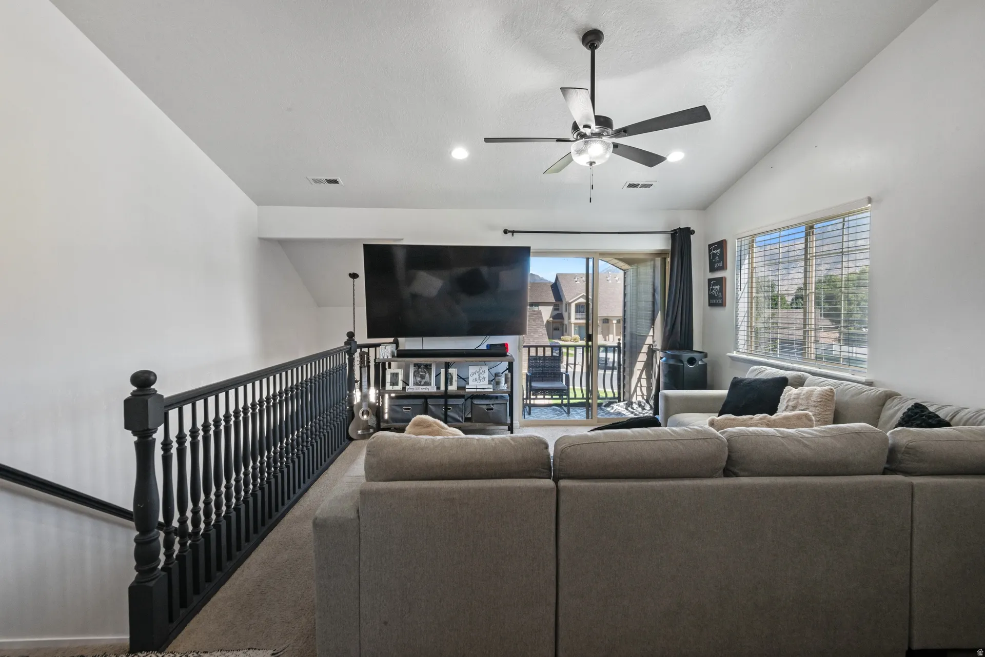 Vaulted ceiling, ceiling fan, large window with blinds, balcony access, and dark wood-finish railing