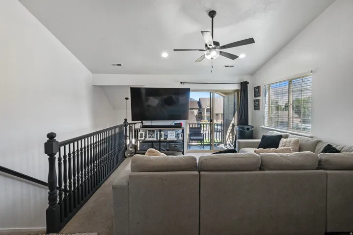 Vaulted ceiling, ceiling fan, large window with blinds, balcony access, and dark wood-finish railing