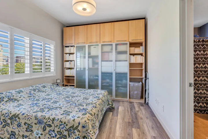 Window with white plantation shutters, wood-finish flooring, and built-in cabinetry with frosted glass panels and open shelving