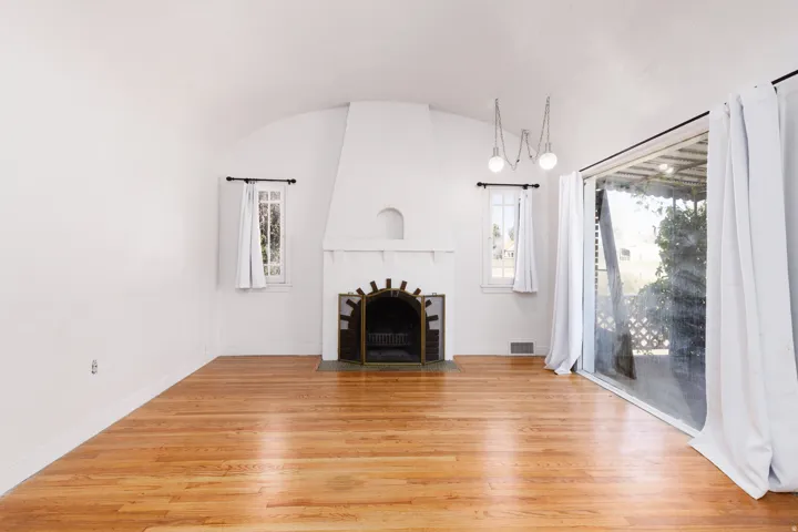 Hardwood flooring throughout, arched ceiling, prominent fireplace with metal screen, two rectangular windows, and a wide sliding glass door