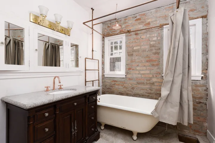 Exposed brick wall, freestanding clawfoot tub, dark wood vanity with white countertop, copper-toned plumbing fixtures, and built-in medicine cabinets